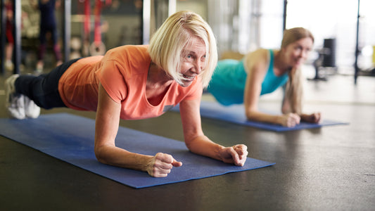 Senior woman practicing plank pose on blue yoga mat wearing orange top and black leggings next to a woman wearing light green top