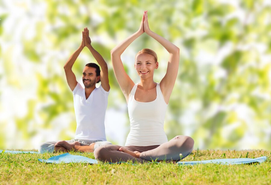 sport-fitness-yoga-people-concept-smiling-couple-meditating-sitting-mats-with-raised-hands-green-tree-leaves-background